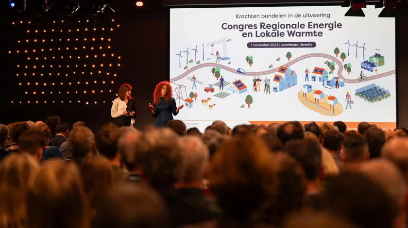 Blik van achteruit de zaal op podium met op het beeldscherm de tekst 'Krachten bundelen in de uitvoering, Congres Regionale Energie en Lokale Warmte, 1 november 2023 | Jaarbeurs, Utrecht' boven een afbeelding van de fysieke leefomgeving.