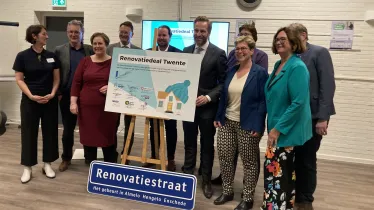 Hugo de Jonge en de vertegenwoordigers van de betrokken gemeenten en woningbouwcorporaties poseren bij een bord met de titel 'Renovatiedeal Twente' waarop de handtekeningen staan. Op de grond staat een straatnaambord met de naam 'Renovatiestraat'.