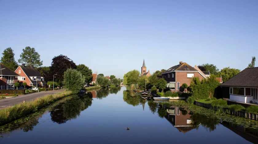 Foto van Bleskensgraaf in Molenland. Kanaal met aan weerszijden vrijstaande en 2-onder-1-kap-woningen en op de achtergrond een kerktoren.