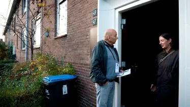 Man met folder in de hand spreekt een vrouw aan die in de deuropening staat