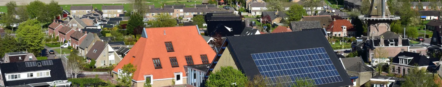 Foto van een oude dorpskern met in het midden een traditioneel Fries boerderijdak met oranje dakpannen en daarnaast een modern zwart puntdak met zonnepanelen. Op de achtergrond een stellingmolen