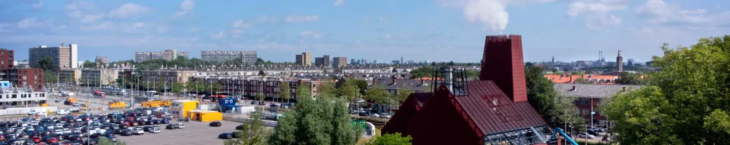 Aardwarmteinstallatie met links een enorme ronde putdeksel en rechts een modern kathedraal-achtig gebouw bekleed met steenrood gecoate stalen roosters. Op de achtergrond links auto's op een parkeerterrein en rechts hoogbouw tegen een blauwe lucht met schapenwolken.