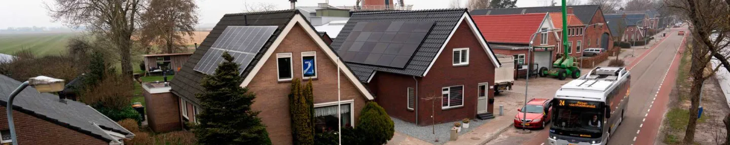 Doorgaande weg in Nieuwe Pekela met aan de linkerkant vrijstaande huizen met puntdaken voorzien van zonnepanelen. Op de weg rijdt bus 24 naar Assen. Op de achtergrond staat een molen.