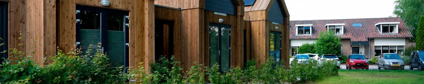 Grasveld met links een drietal houten tiny houses met punttaken voorzien van zonnepanelen. Op de achtergrond jaren '60-rijtjeshuizen.